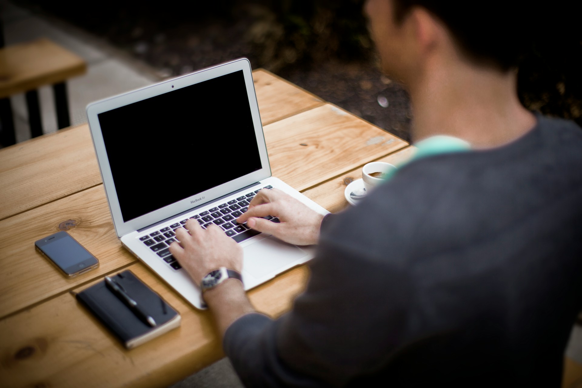 Una persona está sentada en una mesa de madera al aire libre, escribiendo en una computadora portátil. Cerca hay un teléfono inteligente, una computadora portátil y una taza de café. Parecen centrados en escribir contenido SEO amigable para optimizar su sitio web.