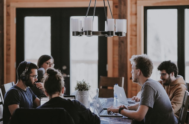 Un grupo de personas sentadas alrededor de una mesa trabajando en computadoras portátiles, dirigiéndose a una "emergencia empresarial", con una moderna lámpara de araña colgando sobre ellos en una habitación con grandes ventanales.