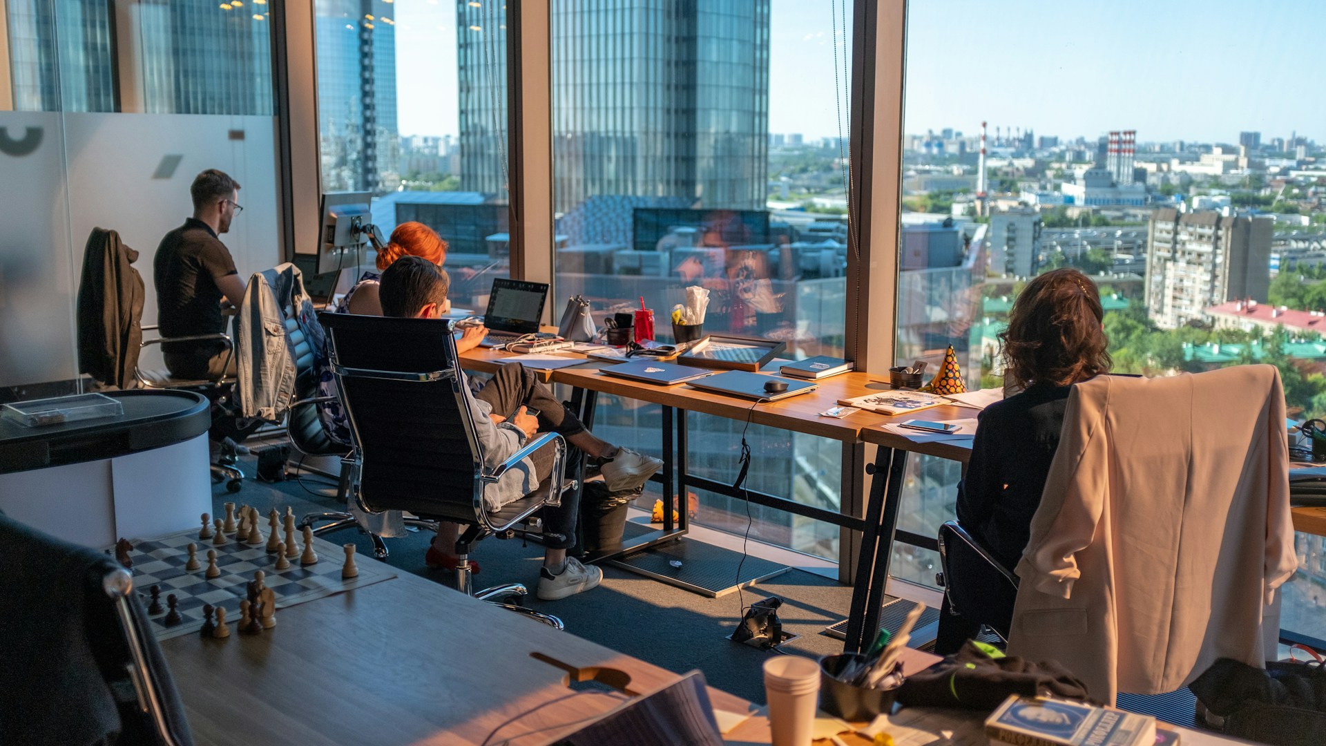 Una oficina con grandes ventanales que muestran una vista de la ciudad; tres personas trabajan en escritorios con computadoras, beneficiándose de la atmósfera tranquila, mientras que un tablero de ajedrez descansa sobre una mesa en primer plano, lo que ilustra el enfoque equilibrado de la agencia hacia los negocios.