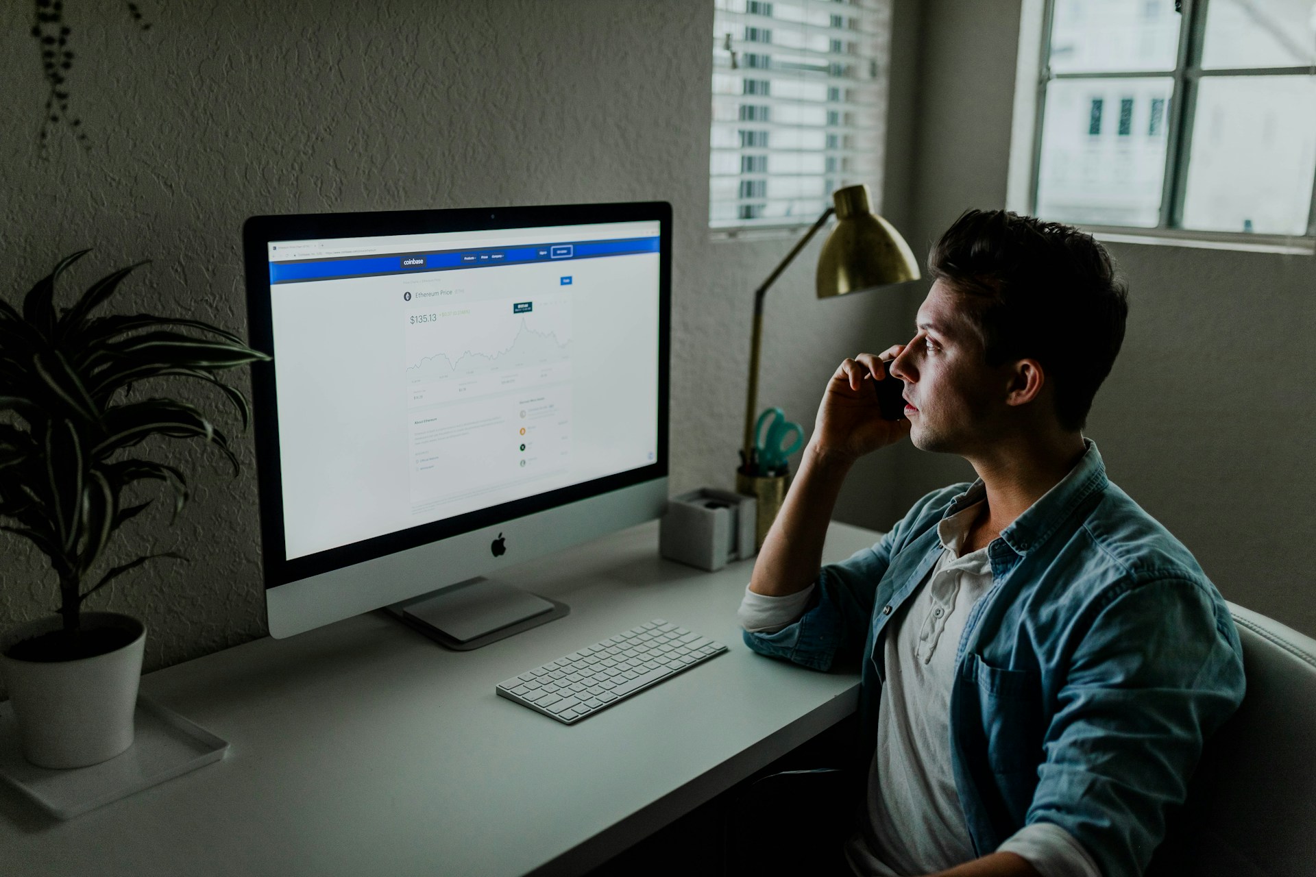 Una persona con una camisa informal está sentada en un escritorio frente a una computadora, hablando por teléfono. La pantalla de la computadora muestra lo que parecen ser gráficos y datos en una página web, probablemente discutiendo la guía definitiva para crear una marca digital efectiva.