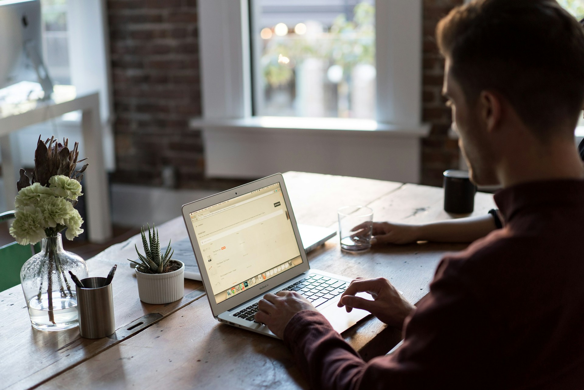 Una persona trabaja en una computadora portátil en una mesa de madera con plantas en macetas, un portalápices y un vaso de agua, tal vez elaborando una publicación interesante para un blog.