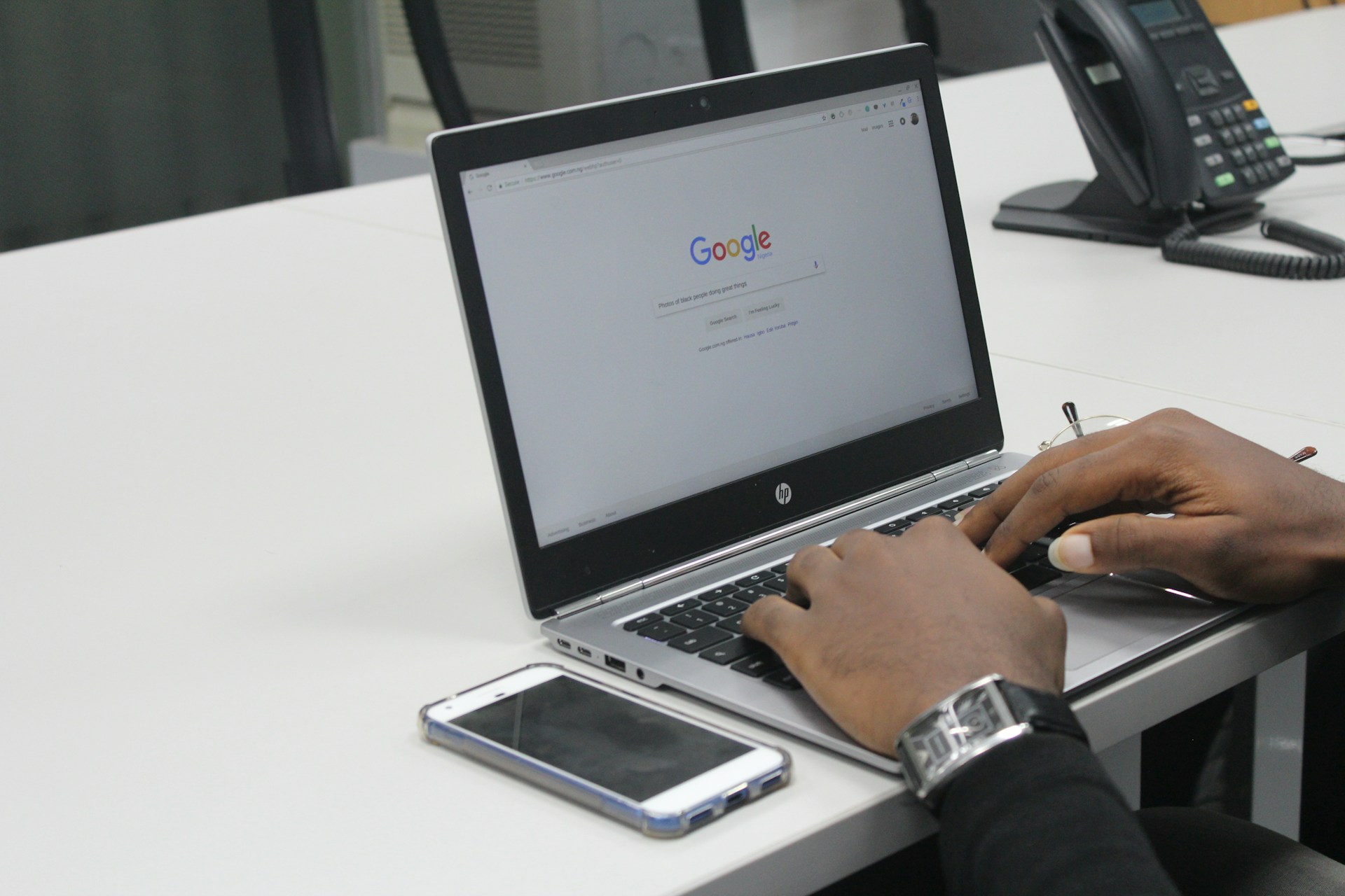 Persona escribiendo en una computadora portátil con la búsqueda de Google abierta, enlaces internos visibles, un teléfono inteligente colocado al lado de la computadora portátil sobre un escritorio blanco y un teléfono al fondo.