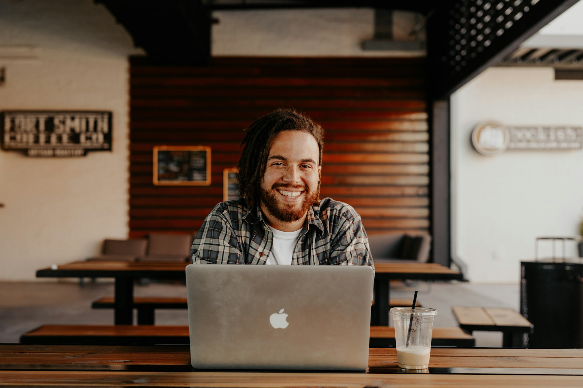 Una persona con rastas sonríe mientras trabaja en una computadora portátil, probablemente concentrada en la actualización de contenido, en una mesa de madera en un espacio interior moderno. Al fondo se ven una bebida y otra mesa.