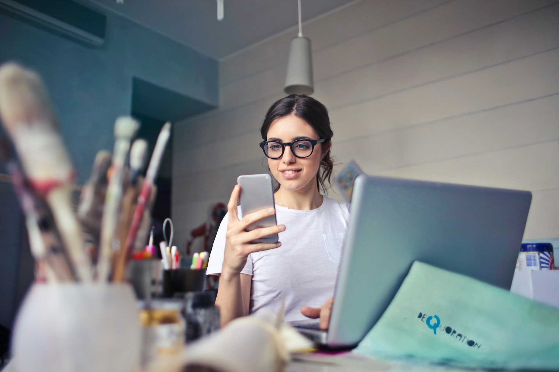 Una mujer con gafas sentada en un escritorio con materiales de arte, mirando su teléfono inteligente, con una computadora portátil abierta frente a ella. Ella administra su tienda en línea de Etsy, optimizando listados de productos con estrategias de SEO para aumentar la visibilidad.