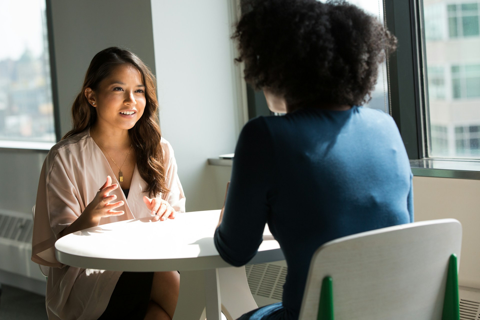 Dos mujeres están sentadas en una mesa redonda blanca cerca de una ventana, conversando dentro de un espacio de oficina bien iluminado. Una mujer hace gestos con las manos mientras habla sobre cómo atraer al público objetivo mediante la creación de contenido que sea atractivo y compatible con SEO.