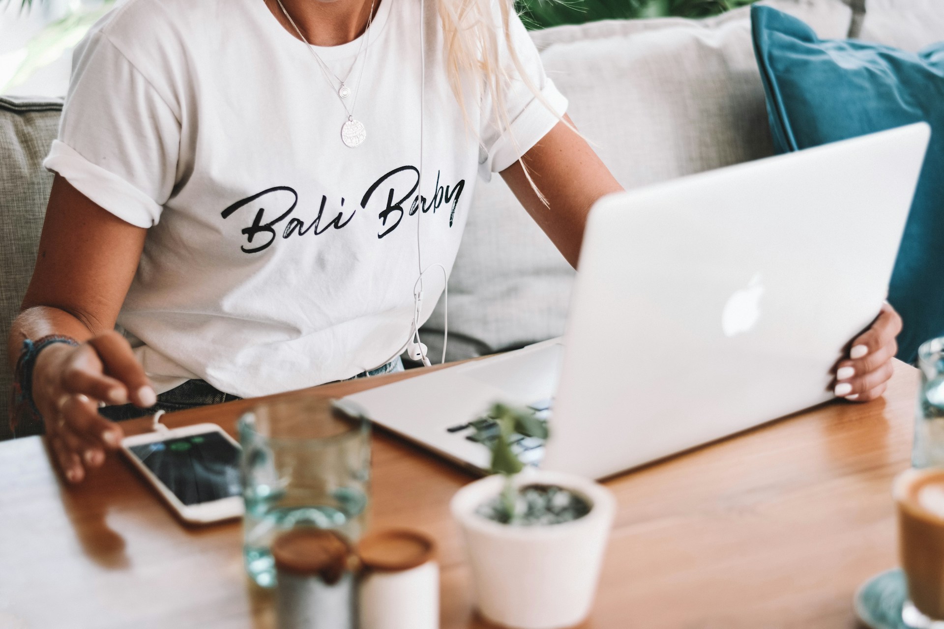 Una persona que lleva una camiseta de "Bali Baby" trabaja en una computadora portátil en una mesa de madera, que también sostiene un teléfono inteligente, un vaso de agua y una pequeña maceta mientras investiga técnicas de construcción de enlaces para mejorar las estrategias de SEO.