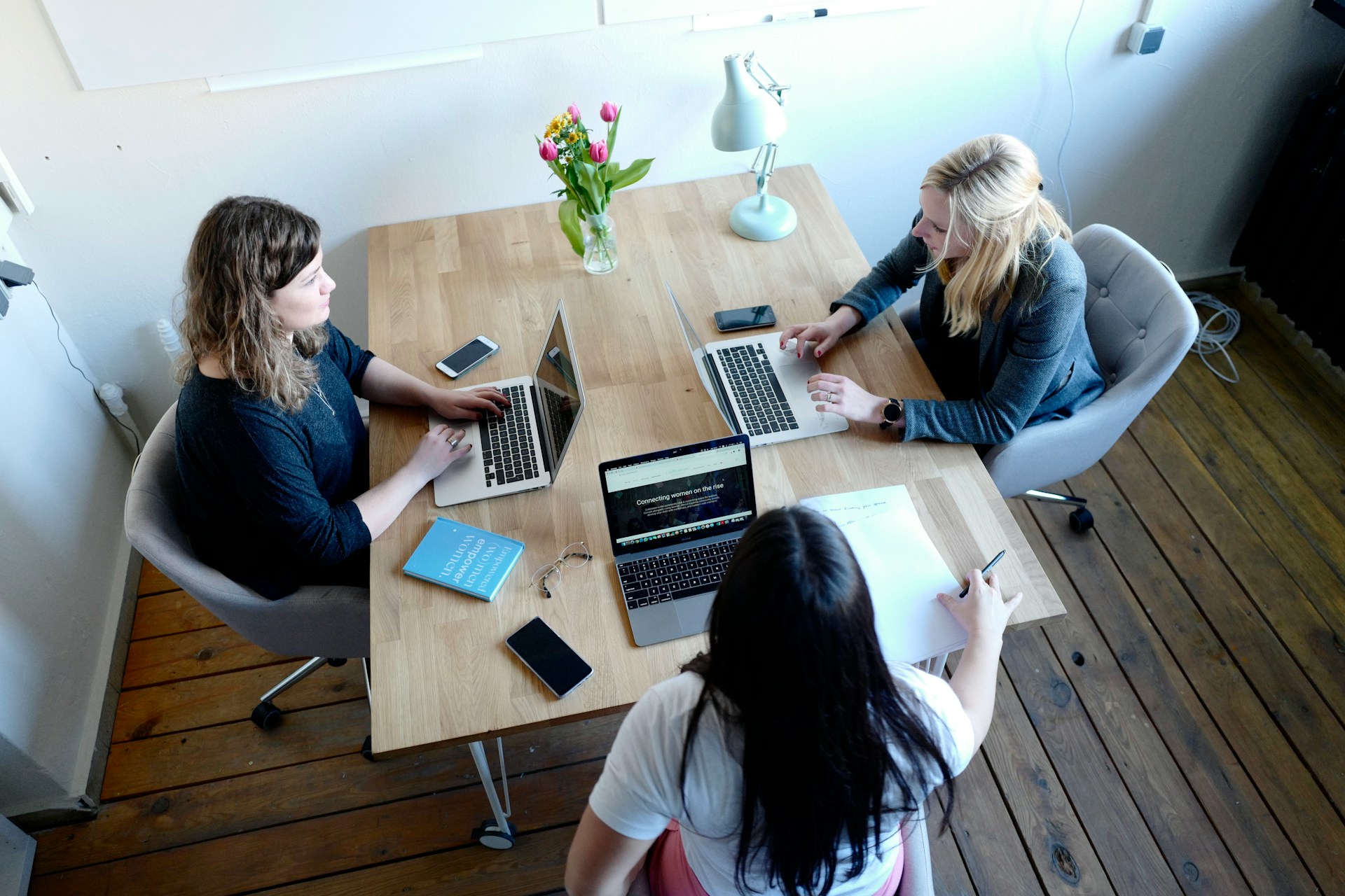 Tres mujeres trabajando juntas en una mesa de madera, cada una usando una computadora portátil. La mesa está adornada con un florero, un libro, un bloc de notas, teléfonos inteligentes y una lámpara de escritorio. Mientras colaboran, su objetivo es mejorar el SEO para elevar la presencia online de su proyecto.