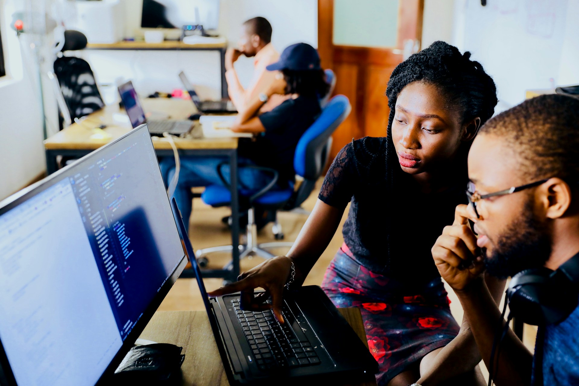 Dos personas trabajan juntas frente a una computadora, centrándose en la pantalla que muestra el código de un sitio web. Al fondo, otras dos personas están sentadas ante escritorios con ordenadores portátiles.