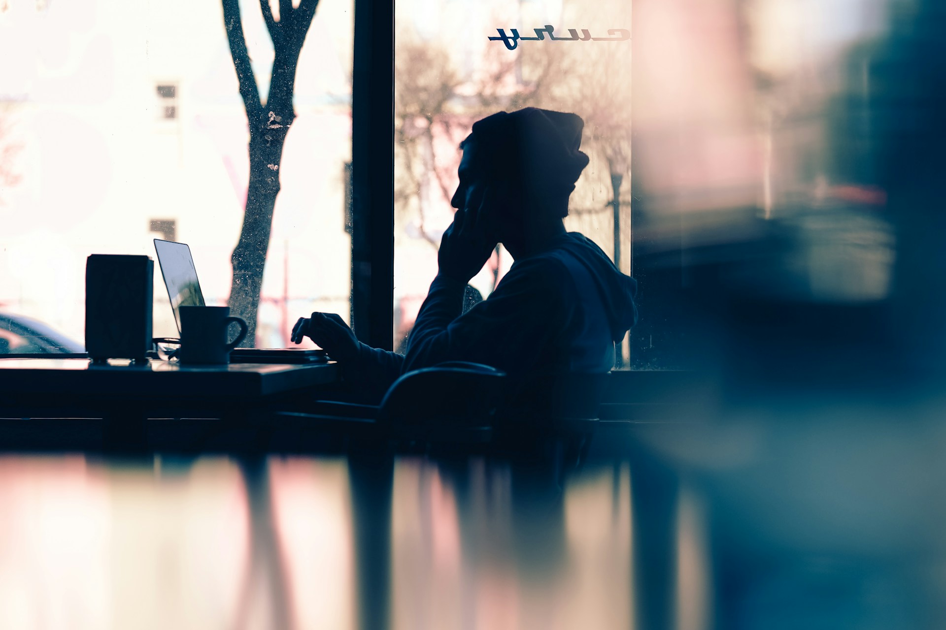 Una persona que lleva un gorro está sentada en una mesa en un café con poca luz, usando una computadora portátil para realizar un análisis SEO mientras sostiene un teléfono junto a su oreja.