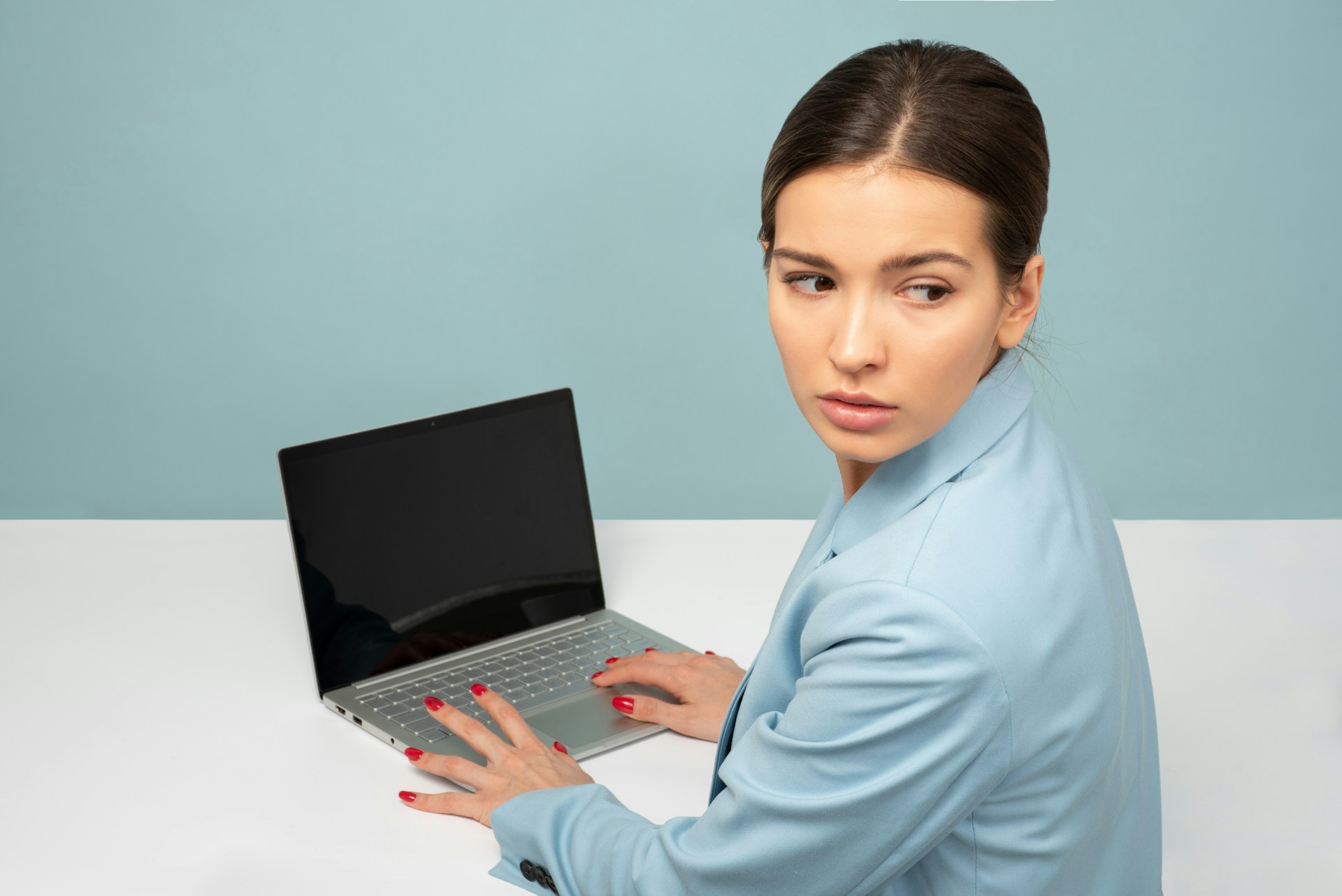 Mujer con una chaqueta azul mirando por encima del hombro mientras usa una computadora portátil en un escritorio blanco con fondo azul, posiblemente revisando errores de SEO.