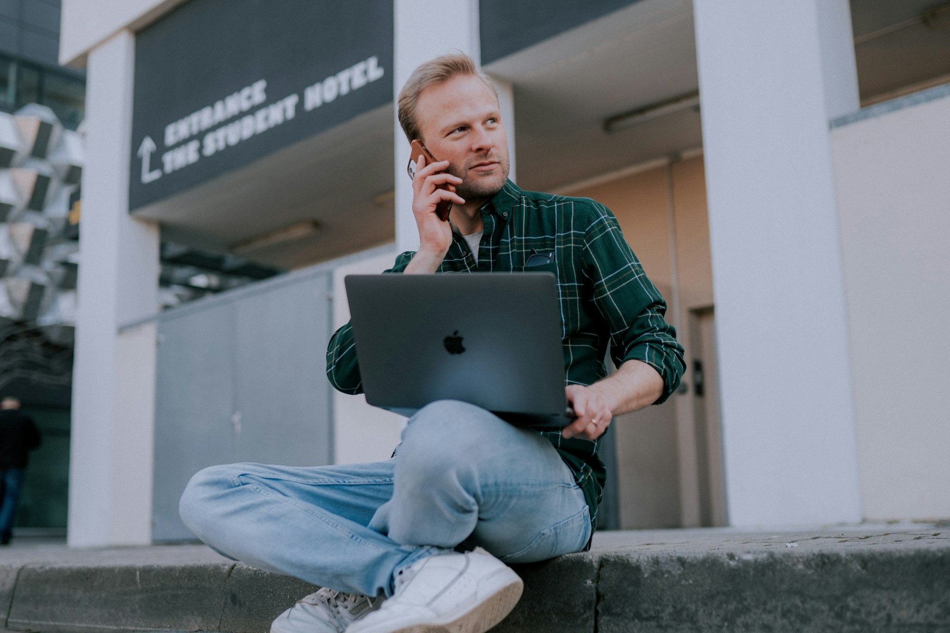 Una persona con una camisa a cuadros verdes y jeans está sentada en el suelo afuera, sosteniendo una computadora portátil y hablando por teléfono cerca de un edificio llamado "The Student Hotel", probablemente discutiendo su negocio en línea.