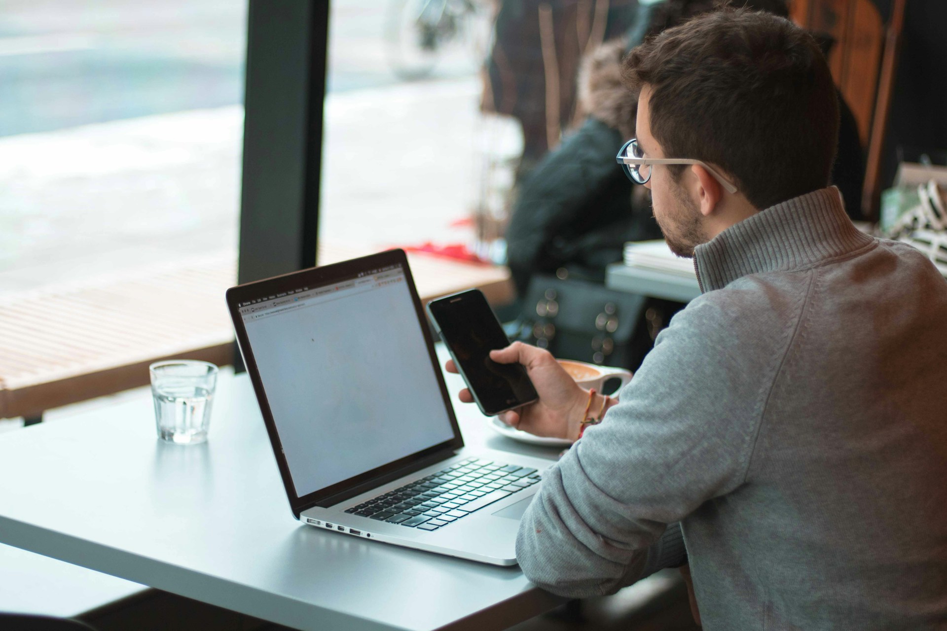 Un hombre con un suéter gris se sienta en una mesa con una computadora portátil y sostiene un teléfono inteligente en una mano, cerca de una ventana de una cafetería. Sobre la mesa hay un vaso de agua y una taza de café mientras contempla la importancia del SEO mientras gestiona su presupuesto de rastreo.