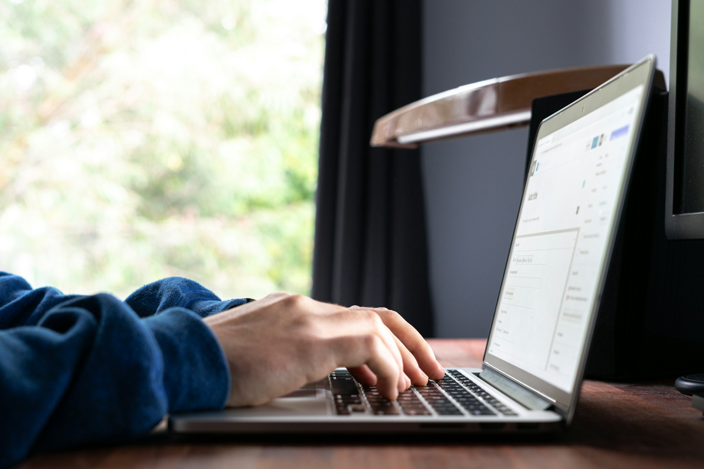 Una persona que lleva un suéter azul escribe en una computadora portátil en un escritorio de madera cerca de una ventana con vista a la vegetación, concentrada en elaborar estrategias efectivas de marketing de contenidos para impulsar el compromiso y las conversiones.