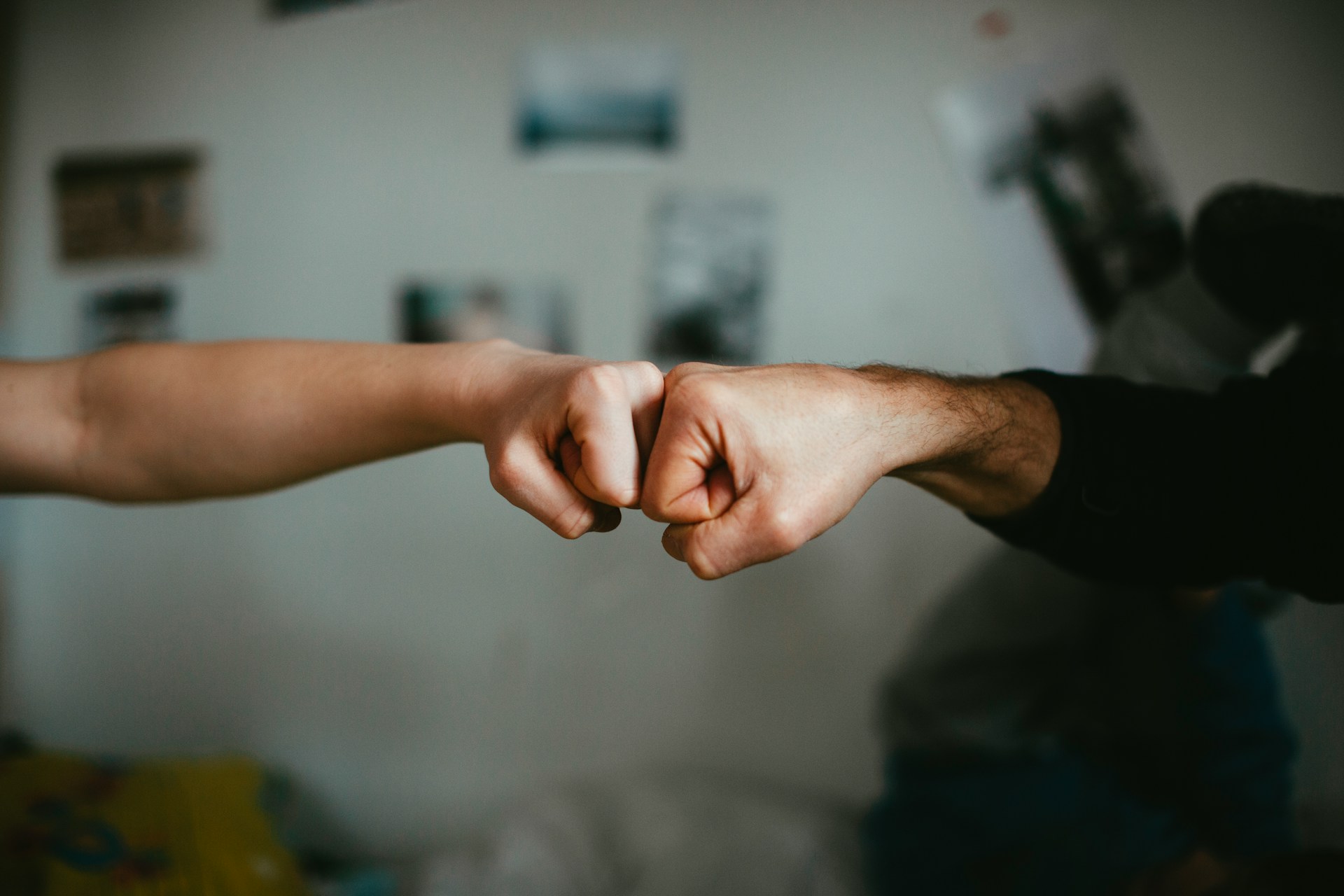 Dos personas chocan los puños en un gesto de amistad o acuerdo, encarnando el espíritu de colaboración crucial para las pymes. El fondo borroso muestra una pared con carteles y fotografías.