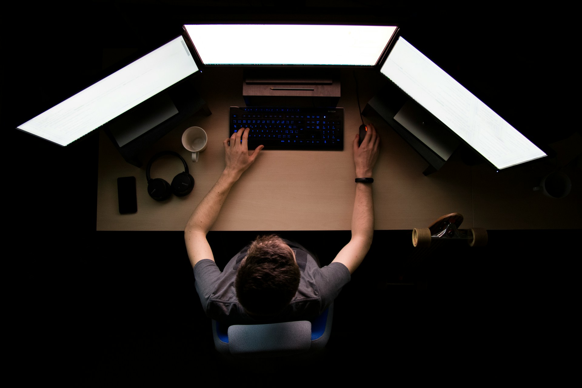 Persona escribiendo en un teclado frente a tres monitores de computadora en una habitación oscura. Sobre el escritorio hay un teléfono, auriculares, una taza de café y una patineta mientras realizan un análisis de las palabras clave del sitio web. Vista de arriba hacia abajo.