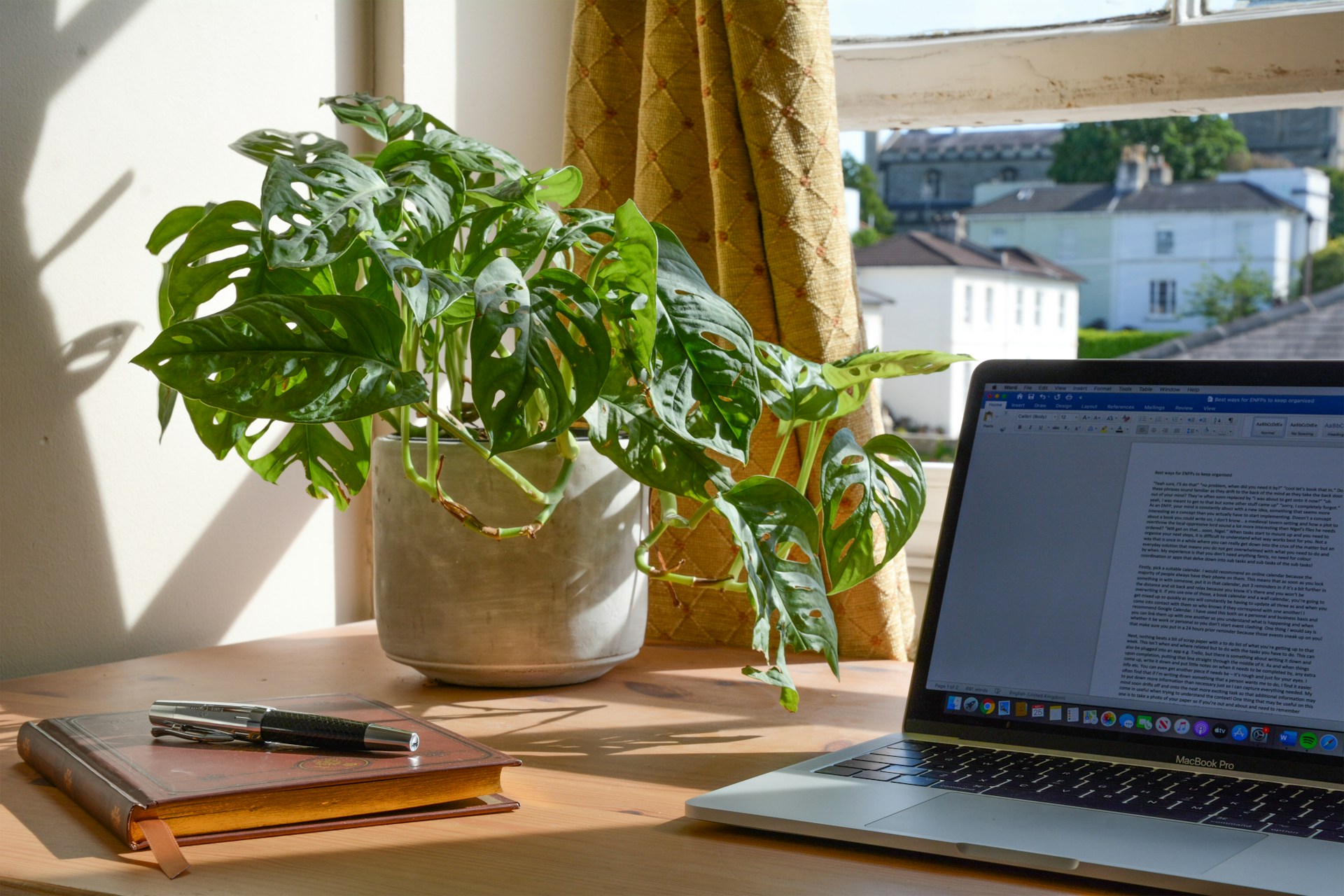 Un escritorio con una computadora portátil abierta que muestra un documento, un cuaderno con dos bolígrafos y una planta en una maceta se encuentran al lado de una ventana con cortinas amarillas. La luz del sol que entra por la ventana realza el escenario perfecto para la creación de contenido de calidad.