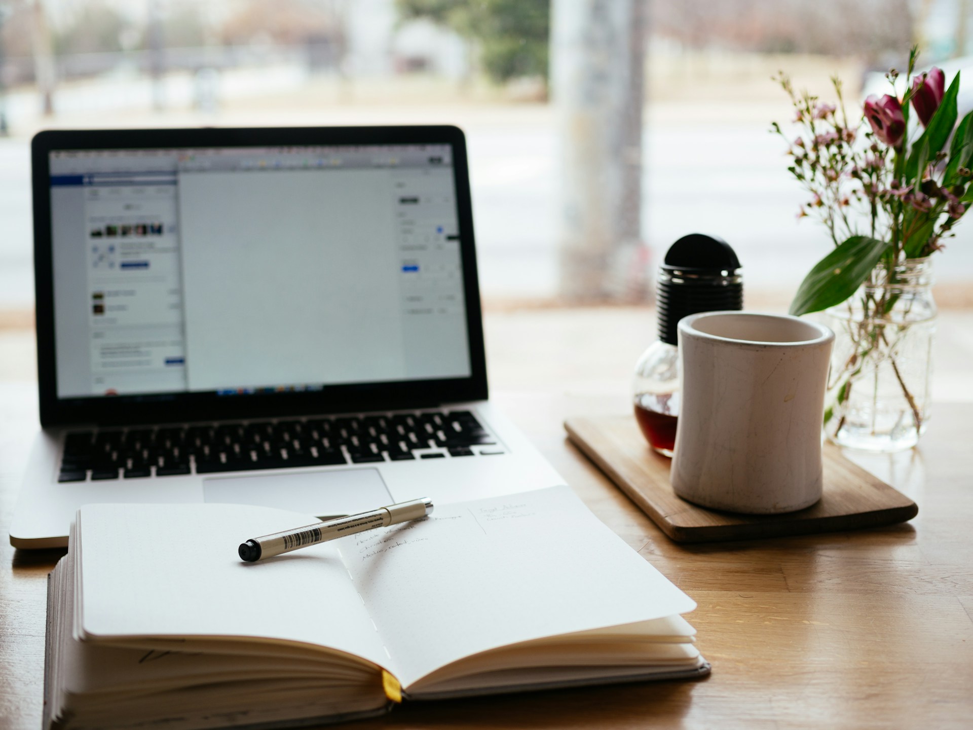 Sobre una mesa de madera cerca de una ventana hay una computadora portátil que muestra un documento sobre estrategia de SEO, una libreta abierta con un bolígrafo, una taza de café, un frasco de vidrio y un jarrón con flores.