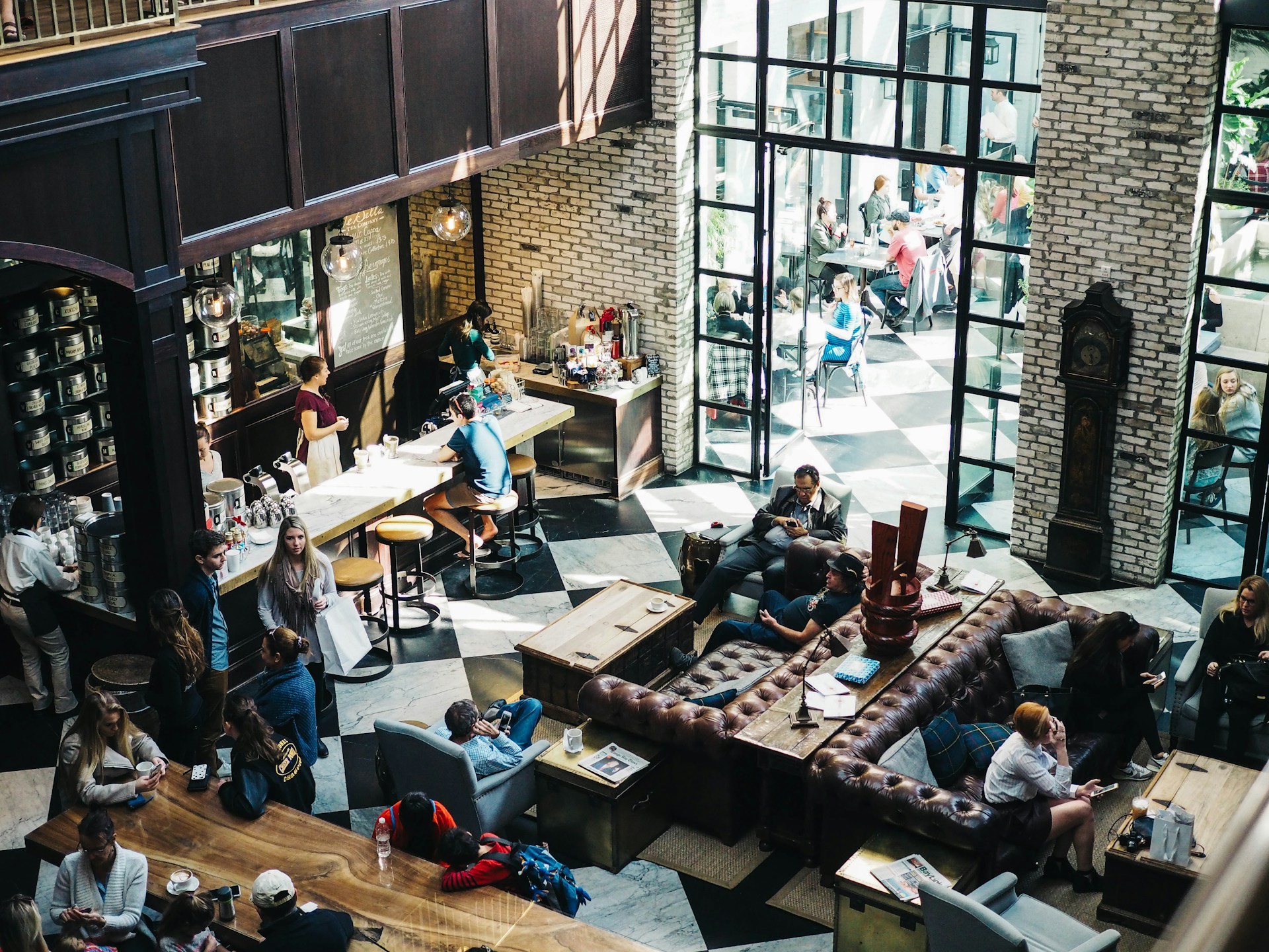 Una cafetería concurrida con gente sentada en mesas, sofás y en la barra del interior, mientras que se ve a otros a través de la puerta de cristal del exterior. Con techos altos y paredes de ladrillo, es un lugar ideal para impulsar el tráfico a tu restaurante utilizando técnicas de SEO.