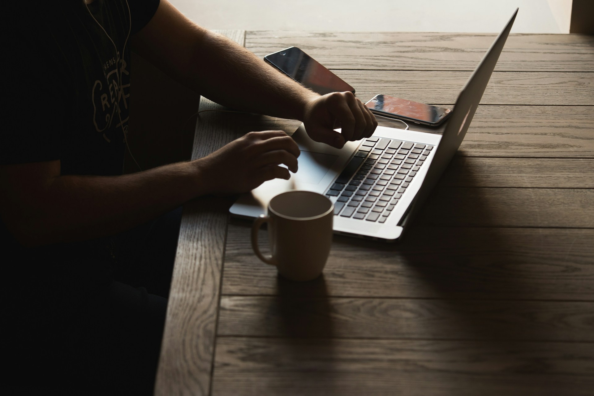 Una persona que trabaja en una computadora portátil en una mesa de madera con una taza de café y dos teléfonos inteligentes, concentrada en optimizar imágenes para mejorar la velocidad de carga del sitio web.