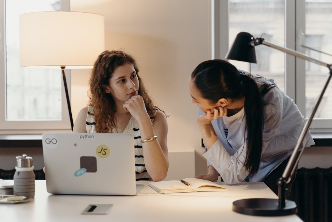 Dos mujeres conversan en un escritorio, una está sentada con una computadora portátil y la otra se inclina sobre una libreta, probablemente discutiendo su análisis de vínculos de retroceso. El escritorio cuenta con una botella de agua, una lámpara y un teléfono.