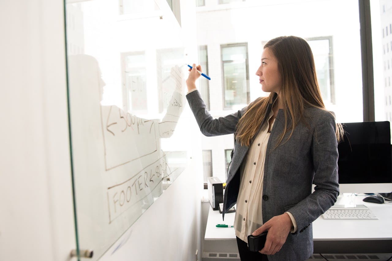 Una mujer vestida de negocios escribe en una pizarra en una oficina bien iluminada, delineando su estrategia comercial, con una computadora y un escritorio visibles al fondo.