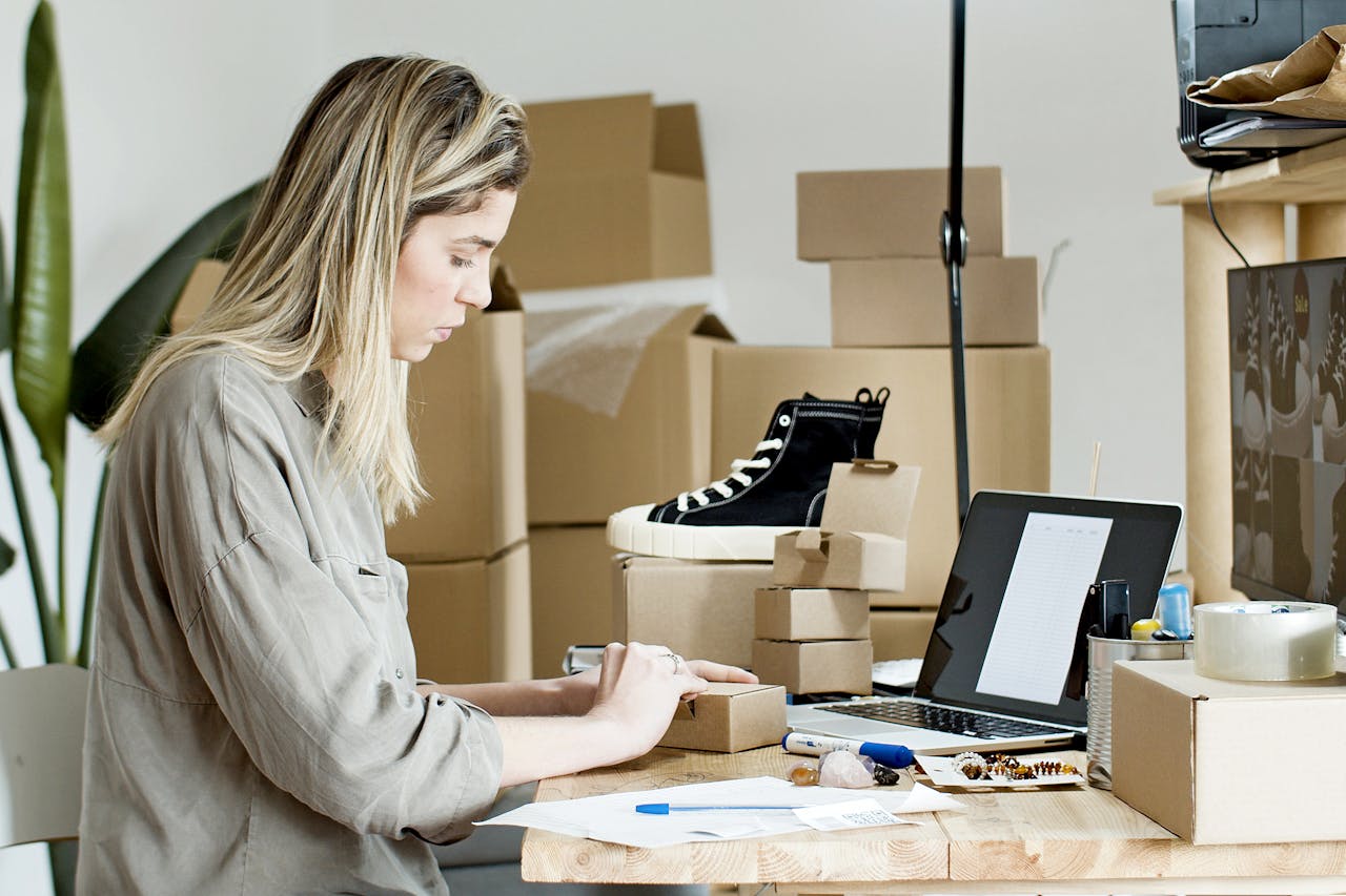 Una mujer está empacando artículos en un escritorio rodeada de cajas de cartón y una computadora portátil, administrando eficientemente su tienda en línea. Sobre el escritorio se ve un único zapato.