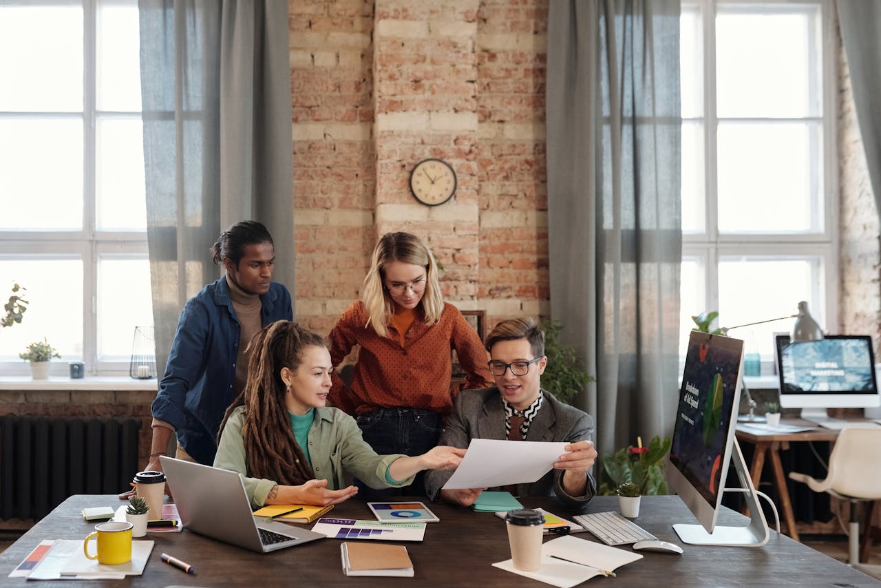 Un grupo de cuatro personas colaborando en un escritorio con portátiles y papeles en una oficina moderna con paredes de ladrillo y grandes ventanales, discutiendo estrategia de marketing digital.