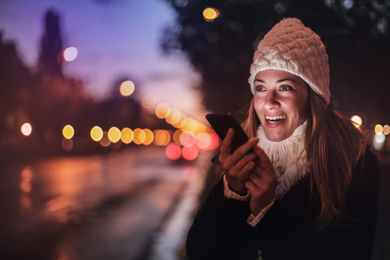 Una mujer está parada al aire libre al anochecer, con un gorro y una bufanda de invierno, sonriendo a la pantalla de su teléfono inteligente. Las farolas desenfocadas crean un efecto bokeh en el fondo, tal vez mientras explora búsquedas por voz o considera Optimización SEO para su nuevo proyecto.