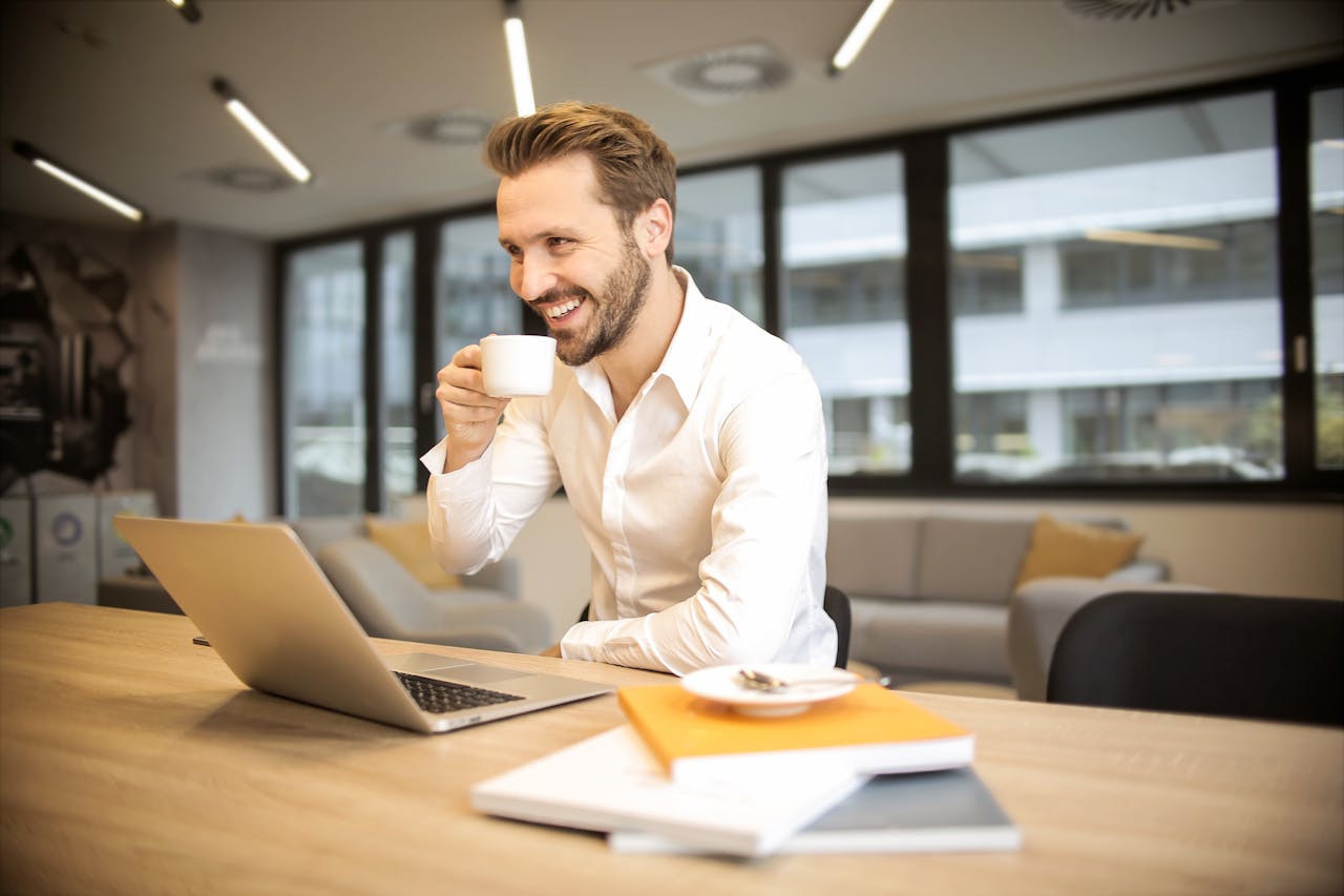 Un hombre está sentado en un escritorio en una oficina, sonriendo y bebiendo de una taza blanca mientras mira su computadora portátil, tal vez optimizando la densidad de palabras clave para su último proyecto.