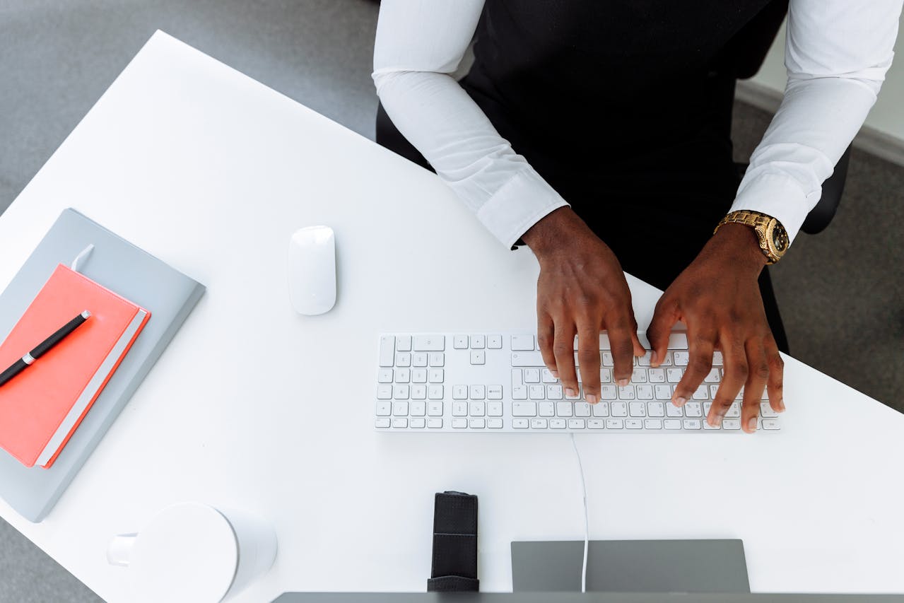 Persona escribiendo en un teclado blanco en un escritorio blanco. Sobre el escritorio hay un cuaderno cerrado con un bolígrafo, un ratón, un smartphone y una taza de café. Con la concentración necesaria para evitar la canibalización de las tareas, la persona viste una camisa blanca y un reloj de oro.