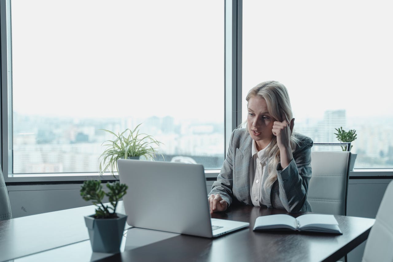 Una mujer con traje de negocios está sentada en un escritorio frente a una computadora portátil, con la mano apoyada en la sien y una libreta abierta a su lado. En esta moderna oficina con grandes ventanales y vista a la ciudad, se enfoca en estrategias para mejorar el SEO y la experiencia del usuario.