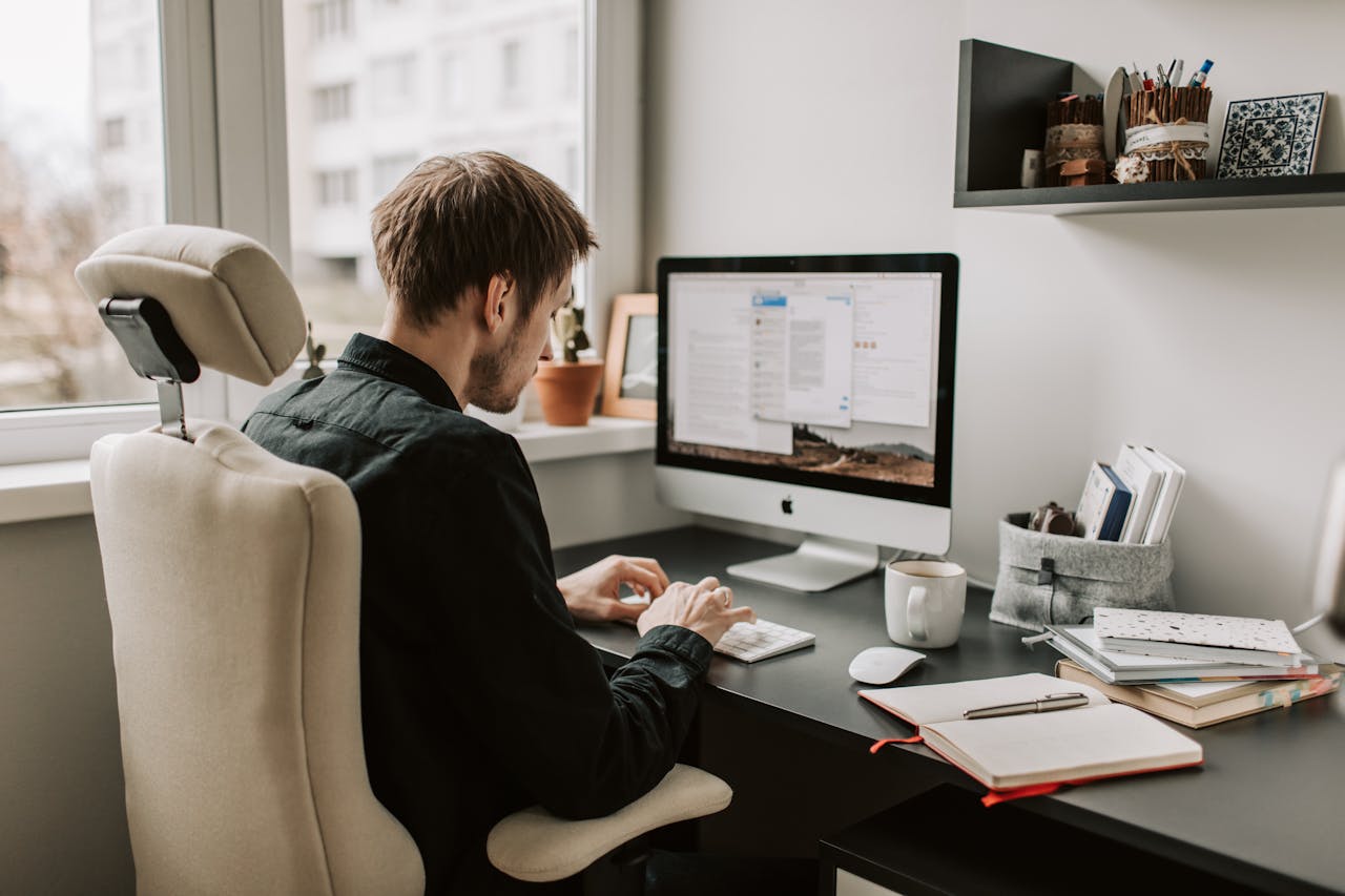 Una persona sentada en un escritorio frente a una computadora, escribiendo en un teclado. El escritorio, adornado con varios elementos como una taza, cuadernos y una planta en maceta, da a entender su enfoque en el marketing de afiliados. En la pared se montan estantes con más elementos.