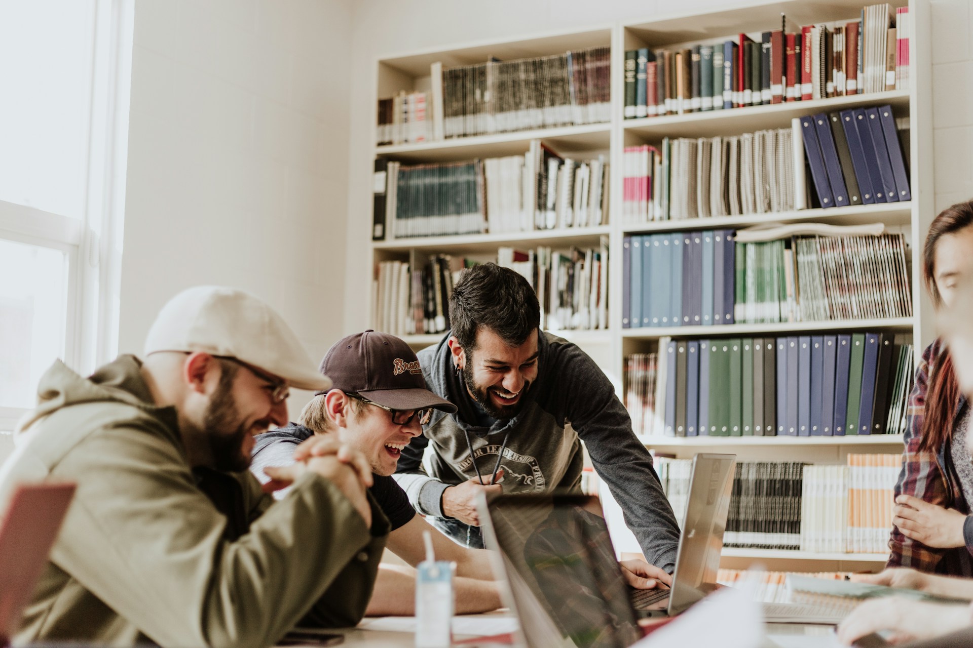 Cuatro personas están reunidas alrededor de una mesa, mirando una computadora portátil y riendo, resaltando la atmósfera alegre de su sesión de lluvia de ideas B2C. Al fondo hay una estantería llena de carpetas y libros de varios colores.