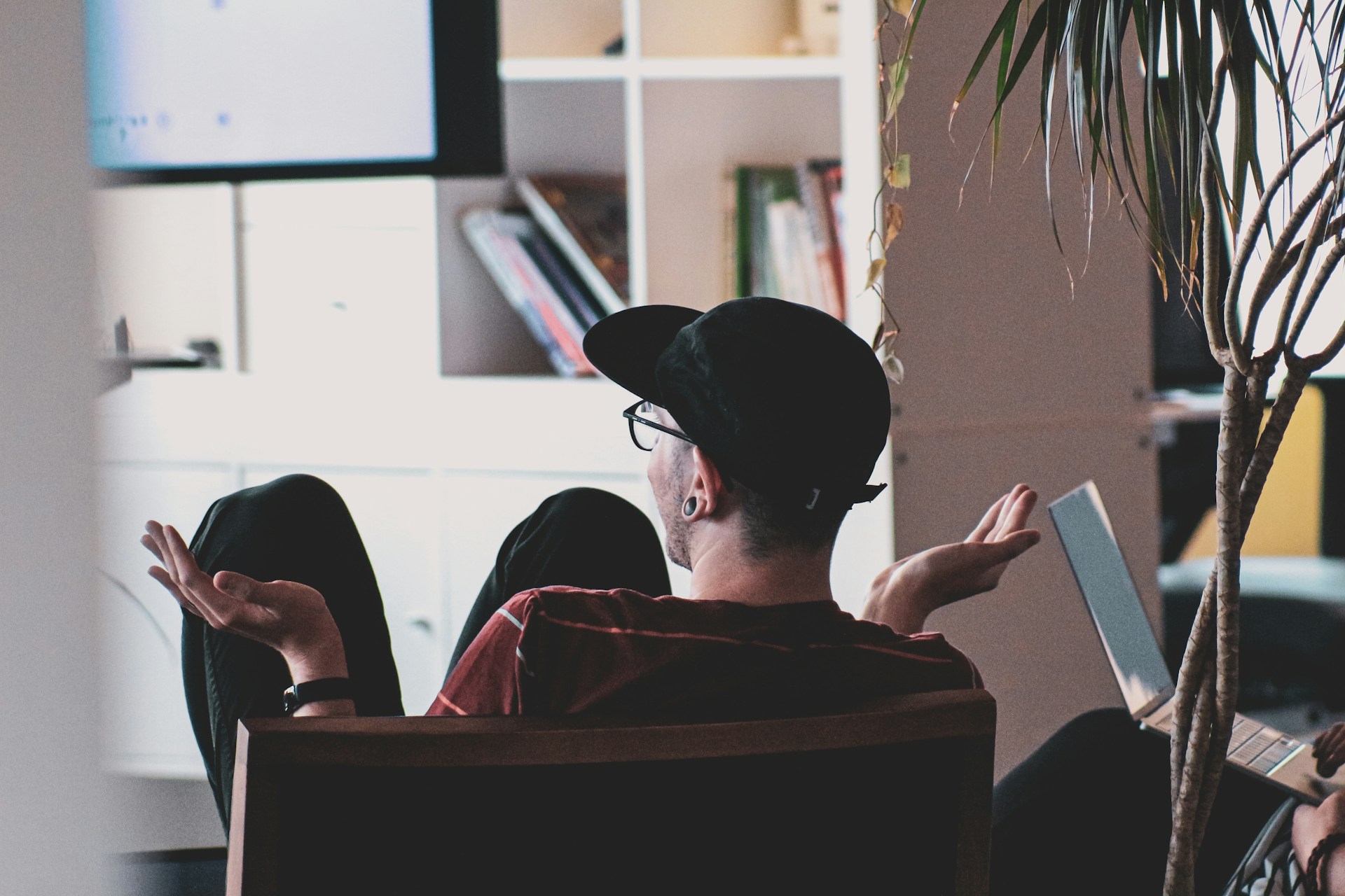 Una persona con gorra se sienta en una silla con los brazos abiertos en gesto interrogativo, mirando una pantalla, tal vez reflexionando sobre SEO para psicólogos. Al fondo hay una planta y estanterías con libros.