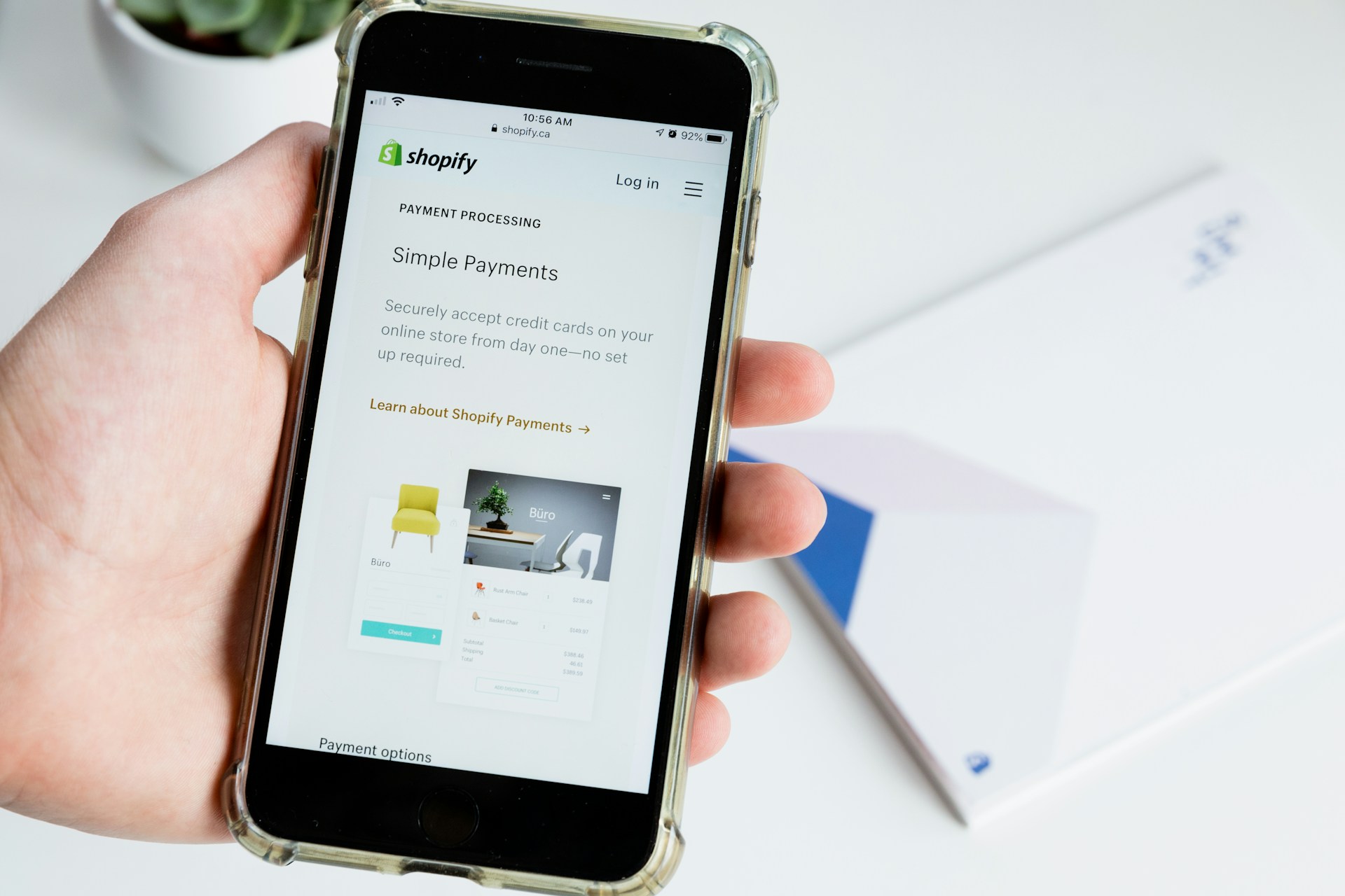 Una mano sosteniendo un teléfono inteligente que muestra una página web de Shopify sobre pagos simples, con un fondo de un escritorio borroso y objetos de una eficiente tienda en línea.