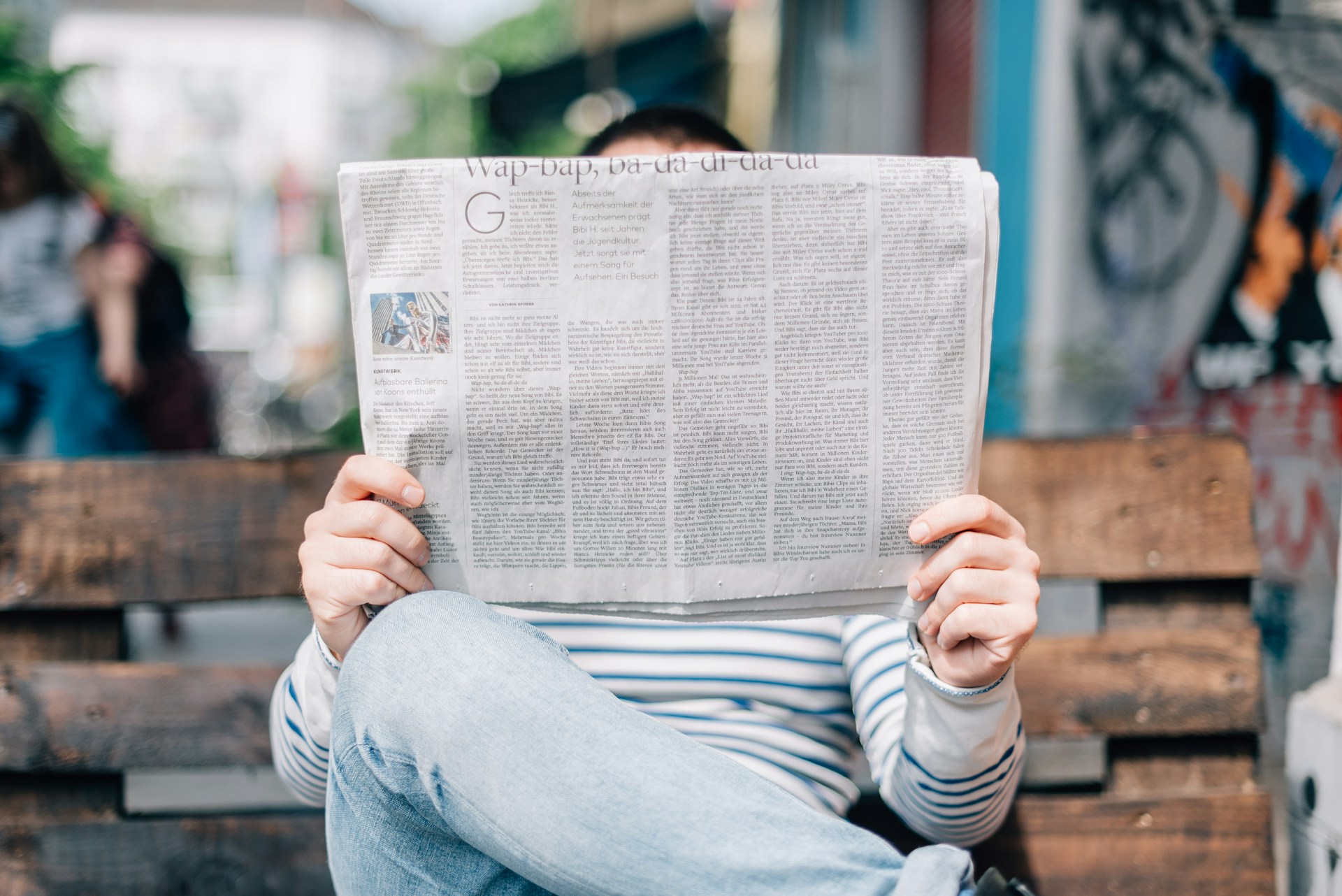 Una persona sentada en un banco lee un periódico, con el rostro oculto detrás de él. Llevan una camisa a rayas y jeans. El fondo es un entorno urbano con detalles borrosos, que se asemeja al telón de fondo de los comunicados de prensa cotidianos.