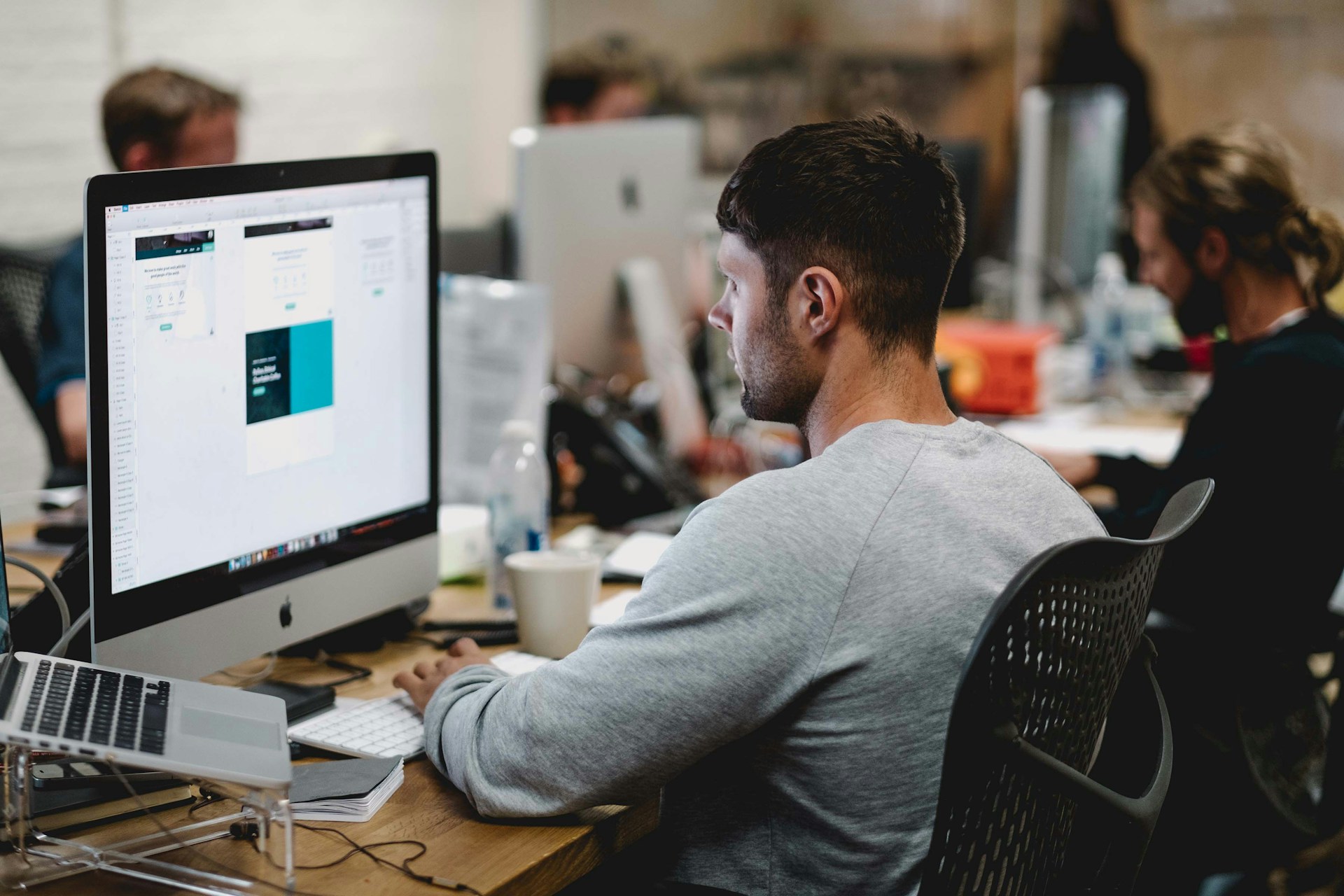 Un hombre con una camisa gris trabajando en una computadora de escritorio en una oficina ocupada, analizando los datos de la tasa de rebote, con varias otras personas y escritorios visibles en el fondo.