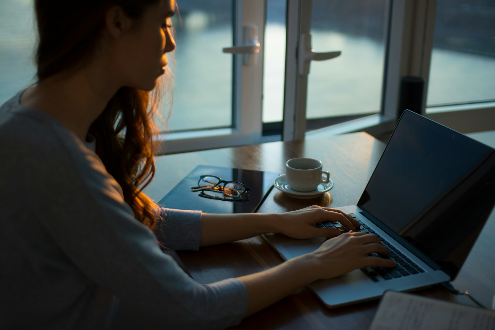 Una persona escribiendo en una computadora portátil en una mesa de madera cerca de una ventana con vista a la ciudad, una taza de café y un par de vasos al lado, profundizando en las complejidades de Web Semántica.