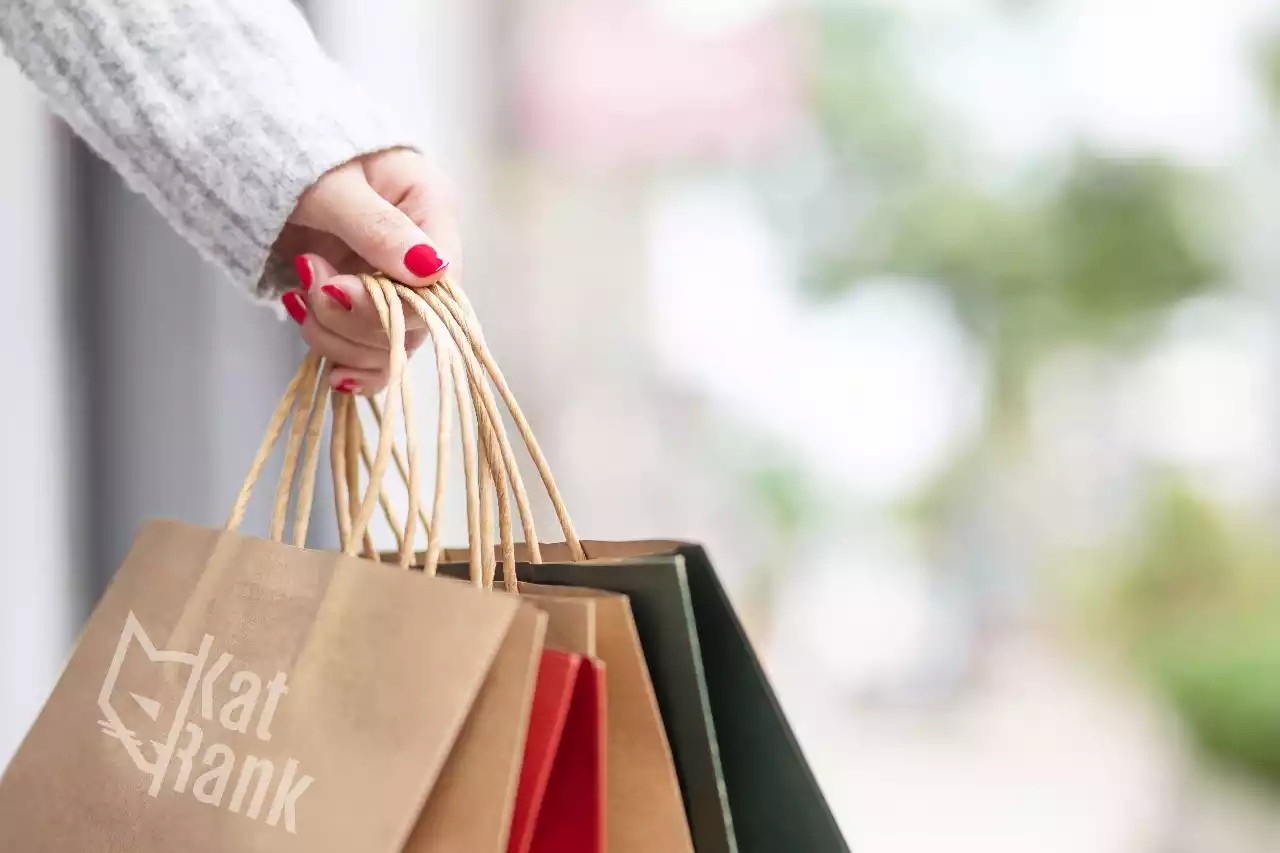 Una persona con esmalte de uñas rojo sostiene varias bolsas de compras de papel marrón y verde, ansiosa por explorar las emocionantes Ofertas de Fin de Año.