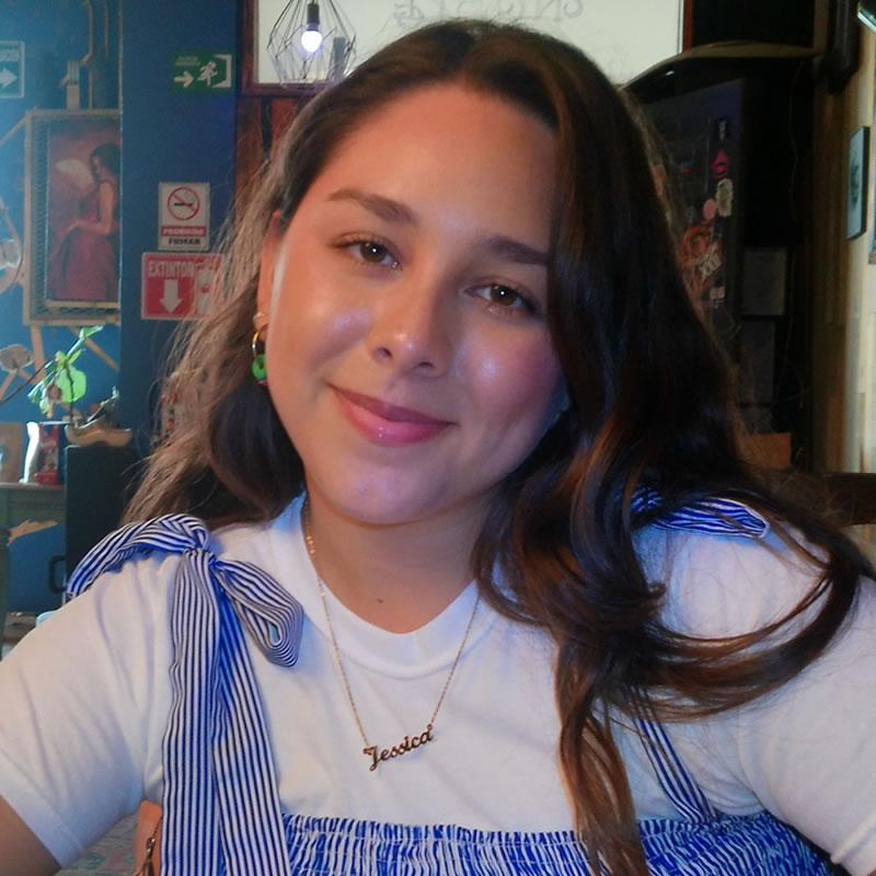 Una mujer vestida de blanco y azul está sentada sonriendo en una mesa en un café acogedor y decorado.